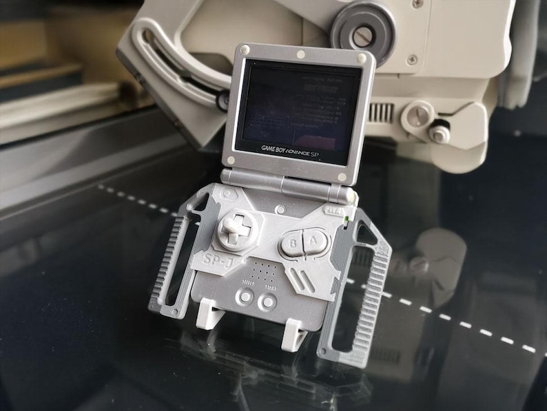 May include: A silver Game Boy Advance SP handheld console with a custom SP-J case. The screen is open, displaying a game. The case has a D-pad, A and B buttons, and the text "SP-J". The console is on a reflective surface.