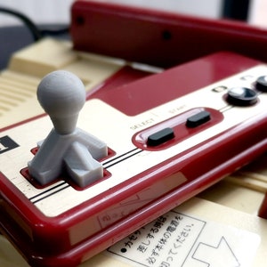Puede incluir: Un mando de control de Nintendo Entertainment System (NES) rojo y blanco con una palanca de mando gris. El mando tiene un botón de selección, un botón de inicio y dos botones redondos. El texto "SELECT" y "START" está impreso en el mando.