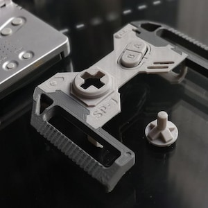 May include: A gray 3D-printed tool with the text "SP-1" and a Game Boy SP-style button layout. The tool has a textured grip and a separate gray button. A silver Game Boy SP is in the background.