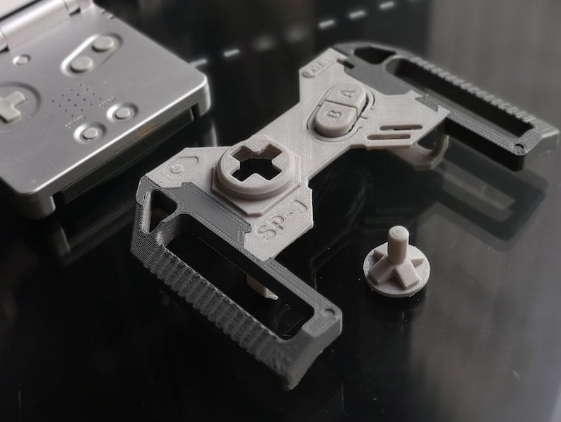 May include: A gray 3D-printed tool with the text "SP-1" and a Game Boy SP-style button layout. The tool has a textured grip and a separate gray button. A silver Game Boy SP is in the background.