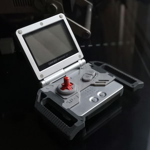 Puede incluir: Consola portátil Game Boy Advance SP plateada, abierta e inclinada. La pantalla es negra y los controles incluyen una cruceta roja y botones grises. El texto "GAME BOY ADVANCE SP" es visible.