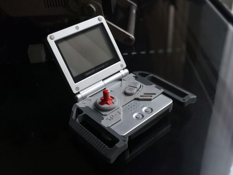 May include: A silver Game Boy Advance SP handheld console, open and angled. The screen is black, and the controls include a red directional pad and gray buttons. The text "GAME BOY ADVANCE SP" is visible.