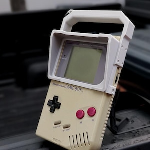 May include: A white and beige Game Boy handheld video game console with a white protective case. The case has a handle and a visor to block out light. The Game Boy has a black D-pad, two pink buttons, and a screen with a grey border.