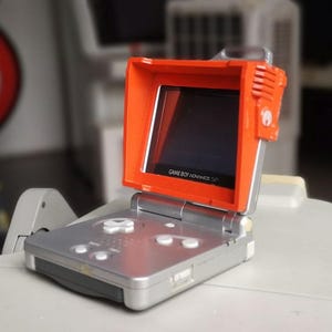 Könnte beinhalten: Ein silbernes Game Boy Advance SP Handheld-Spielkonsolen mit einem orangefarbenen Bildschirmvisier. Der Bildschirm zeigt den Text "GAME BOY ADVANCE SP". Die Konsole hat weiße Tasten und ein Steuerkreuz.