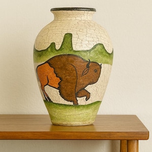 May include: A decorative ceramic vase with a hand-painted bison design. The vase has a cream-coloured background with a cracked texture, a brown bison, and green and brown landscape elements. The rim is black.