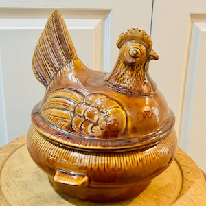 May include: A brown ceramic chicken-shaped container with a lid. The container has a glossy finish and detailed feather and comb textures. The base is round with vertical ridges, and the lid forms the chicken's body and head.