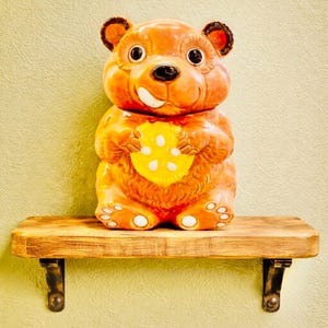 May include: A ceramic bear-shaped cookie jar in shades of orange and brown, holding a yellow cheese wedge. The jar has a smiling face and is displayed on a wooden shelf with metal brackets, against a light yellow wall.