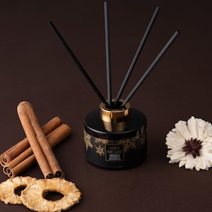 May include: A black glass bottle with a gold lid and black diffuser sticks. The bottle has a gold label with the text "LORIS NICHE AROMATIC CINNAMON Reed Diffuser". The bottle is surrounded by cinnamon sticks and dried pineapple slices.