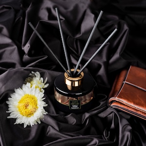May include: A black glass bottle with gold accents and black reeds, filled with a reed diffuser. The bottle has a label that reads "LORIS NICHE BLACK LEATHER Reed Diffuser". The bottle is sitting on a black satin fabric with white flowers.