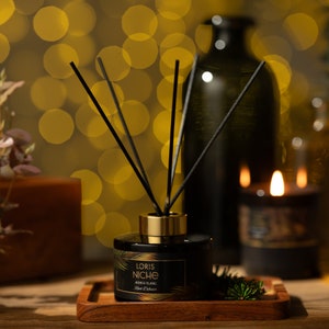 May include: A black glass reed diffuser bottle with gold accents and a label that reads "Loris Niche, Aoud & Ylang, Reed Diffuser". The bottle is surrounded by black reeds and sits on a wooden tray.