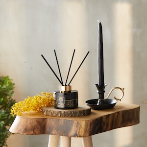 May include: Black and gold diffuser bottle with black reeds and a black candle holder with a gold handle on a wooden table.
