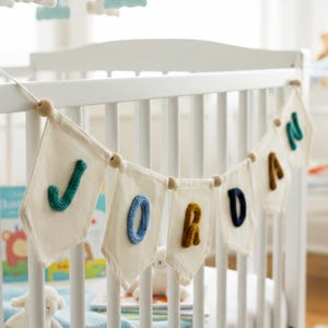 May include: A white crib with a banner spelling "JORDAN" in colorful, textured letters. The letters are attached to small white flags. A mobile with clouds and stars hangs above the crib. The room has a soft, neutral color palette.