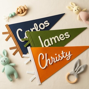 Puede incluir: Tres banderines de fieltro en azul marino, verde y naranja, cada uno con un nombre en letras de cuerda blanca: Carlos, James y Christy. Las banderas tienen adornos decorativos y están rodeadas de juguetes para bebés.