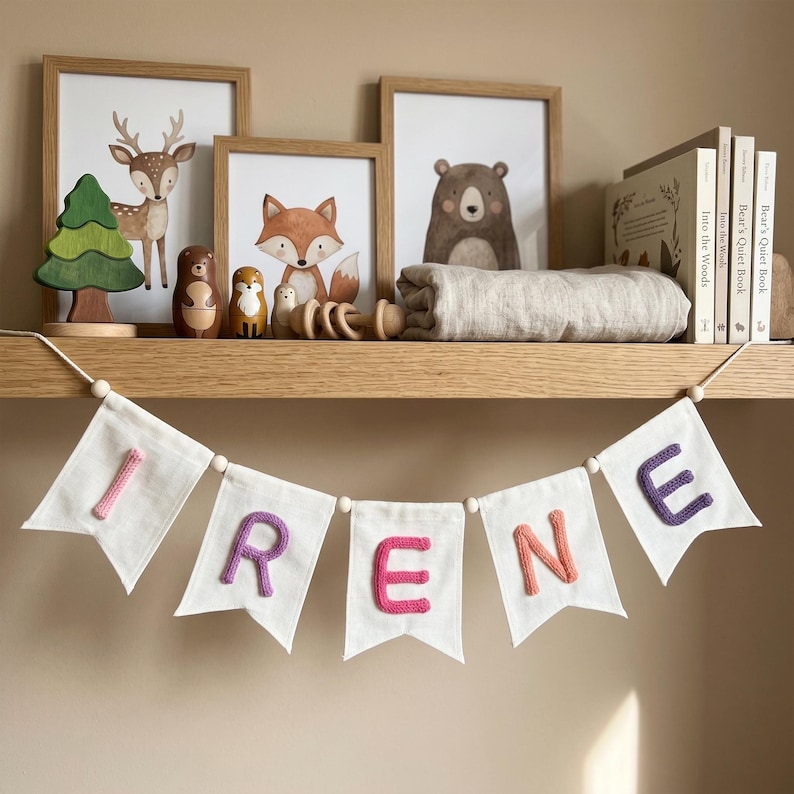 May include: A white fabric banner featuring the name IRENE in colorful, textured, knitted letters. Each letter is centered on a separate pennant flag, connected by a string with small wooden beads between each flag, hanging from a wooden shelf.