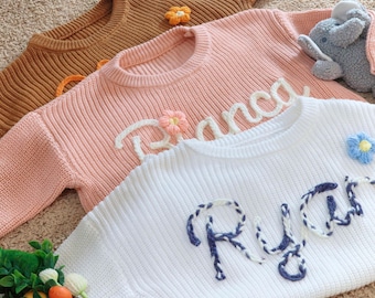 May include: Three knitted sweaters with embroidered names. The top sweater is brown, the middle sweater is pink with the name "Bianca" embroidered in white, and the bottom sweater is white with the name "Ryan" embroidered in blue.