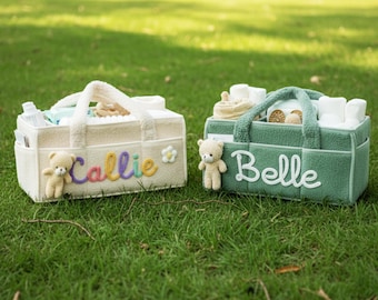 May include: Two plush baby caddies sit on green grass. One is cream-colored with the name "Callie" in rainbow embroidery and a teddy bear. The other is green with the name "Belle" and a teddy bear.