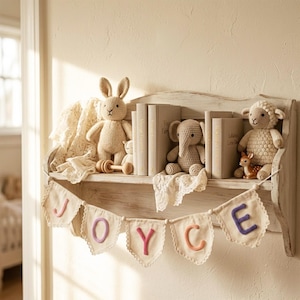 May include: A distressed white wooden wall shelf holding three books, a crocheted beige rabbit, a crocheted gray elephant, a crocheted white sheep, and a small deer figurine. A fabric bunting banner with the letters J-O-Y-C-E hangs from the shelf.