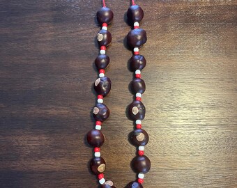 May include: A handmade necklace featuring large, dark brown beads, interspersed with small red and white beads. The necklace is strung on a red cord and is displayed against a dark wood surface. The beads appear to be natural.