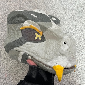 May include: A gray and black snake mask with yellow accents. The mask has a textured surface and a mesh eye covering with a yellow cross design. The snake's mouth features yellow fangs.