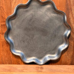 May include: A circular metal tray with a dark gray finish and a wavy, scalloped edge. The surface of the tray has a smooth, matte texture with subtle variations in the metallic tone, creating a simple and functional design for holding items.