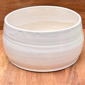 Puede incluir: Un cuenco de cerámica ancho y circular con un esmalte suave de color blanco roto. El exterior presenta sutiles crestas horizontales que rodean el cuerpo, mientras que el interior es profundo y abierto con un acabado blanco limpio y uniforme.