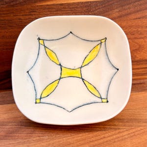May include: A square ceramic dish with rounded corners featuring a white base and a central graphic design. The pattern consists of thin black lines forming a geometric shape with four yellow almond-shaped accents and a central yellow square.