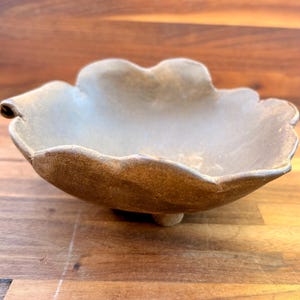 May include: A shallow, hand-formed ceramic bowl with an irregular, wavy rim. The exterior features a textured, earthy brown finish, while the interior has a smooth, light beige glaze. The bowl is supported by a small, central base.