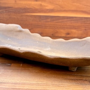 May include: An elongated, shallow ceramic tray with a matte, earthy brown finish. The tray features a scalloped, wavy rim and is supported by small, integrated feet at the base, creating a low-profile, organic, and textured appearance.