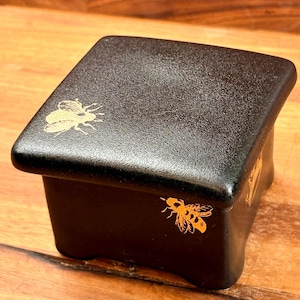 May include: A square black box with a lid, featuring a matte finish and gold-colored bee illustrations on the top surface and the front side. The box has slightly curved sides and a smooth, uniform texture across its dark exterior surface.