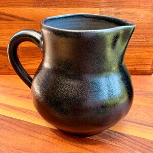 May include: A ceramic pitcher with a rounded body and a curved handle. The exterior features a dark, speckled black glaze with a subtle metallic sheen, while the interior is finished in a smooth, light gray tone. The rim includes a small spout.