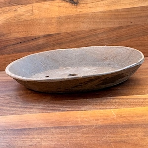 May include: An oval-shaped, shallow gray ceramic bonsai pot with a textured, matte finish. The interior base features two small drainage holes. The vessel has a simple, low-profile design with slightly tapered sides and a smooth, rounded rim.