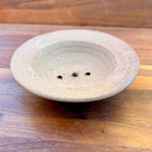 May include: A round, beige ceramic soap dish with a wide, flat rim and a concave center. The base of the dish features three small, circular drainage holes arranged in a horizontal line, with a speckled, textured finish throughout the surface.
