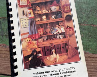 May include: A spiral-bound cookbook titled Making the Aviary a Reality The Court Manor Cookbook Court Manor Nursing Home Ashland, Wisconsin. The cover features a photograph of a wooden hutch filled with decorative items, teddy bears, and geese.