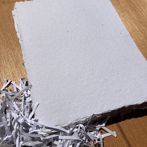 May include: A white handmade paper sheet with a rough, textured surface. The paper is lying on a wooden surface with shredded paper in the foreground.