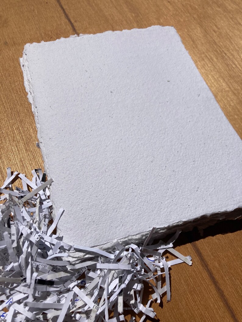 May include: A white handmade paper sheet with a rough, textured surface. The paper is lying on a wooden surface with shredded paper in the foreground.