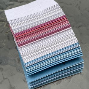 Puede incluir: Una pila de hojas de papel hechas a mano en varios colores. La secci&oacute;n superior es blanca y gris, seguida de una banda de rosa y rojo, y la secci&oacute;n inferior es azul. El papel tiene un aspecto texturizado.