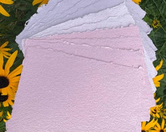 May include: A stack of square sheets of handmade paper with rough, deckled edges. The paper sheets are arranged in a gradient of colors, transitioning from a pale, off-white shade at the top to a soft, light pink hue at the bottom of the stack.