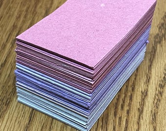 May include: A stack of rectangular cards in shades of pink, purple, and blue. The cards are stacked neatly, with the top card a solid pink color. The edges of the cards are visible, showing the different colors. The cards are on a wooden surface.