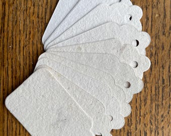 May include: A fan of blank, off-white paper tags with scalloped edges and a hole for stringing. The tags are arranged on a wooden surface. The tags are a neutral color.