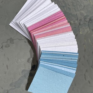 Puede incluir: Un abanico de peque&ntilde;as tarjetas de papel cuadradas en tonos blanco, rosa y azul. Las tarjetas est&aacute;n dispuestas en forma curva, con los colores que van de claro a oscuro. El papel tiene un aspecto texturizado.