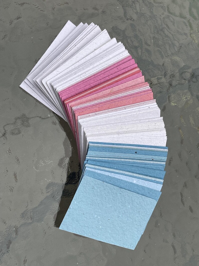 Puede incluir: Un abanico de peque&ntilde;as tarjetas de papel cuadradas en tonos blanco, rosa y azul. Las tarjetas est&aacute;n dispuestas en forma curva, con los colores que van de claro a oscuro. El papel tiene un aspecto texturizado.