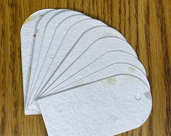 May include: A stack of blank, off-white paper tags with rounded tops and a small hole at the top for stringing. The tags are arranged in a fan shape, resting on a wooden surface.
