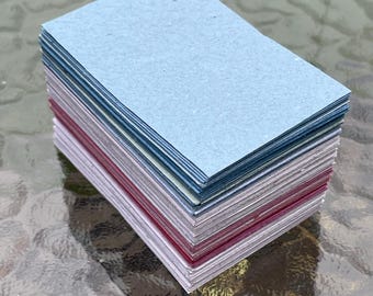 May include: A stack of rectangular cards in various colors, including light blue, silver, and burgundy. The cards are neatly stacked, with the edges visible, creating a layered effect. The cards are on a glass surface.