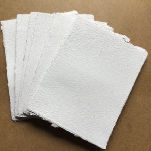 May include: A stack of white handmade paper sheets with a rough, textured surface. The paper is arranged in a fan shape and has a slightly uneven edge.