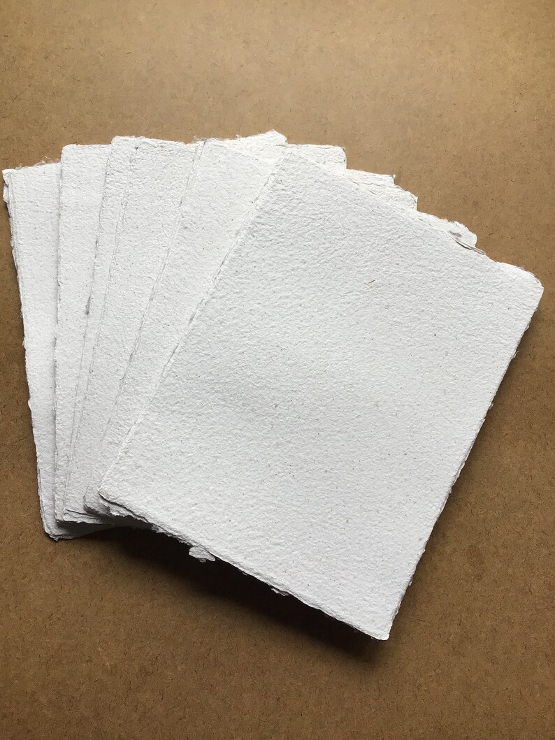 May include: A stack of white handmade paper sheets with a rough, textured surface. The paper is arranged in a fan shape and has a slightly uneven edge.