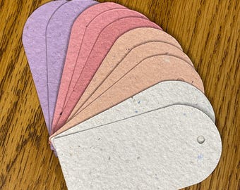 May include: A fan of colorful, textured paper tags in shades of lavender, pink, peach, and off-white. Each tag has a rounded top and a hole for stringing. The tags are arranged on a wooden surface.
