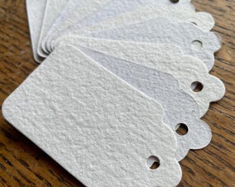 May include: A stack of white, textured paper tags with scalloped edges and a small hole at the top for stringing. The tags are arranged in a fan shape, with the top edges visible. The tags are blank and ready for use.