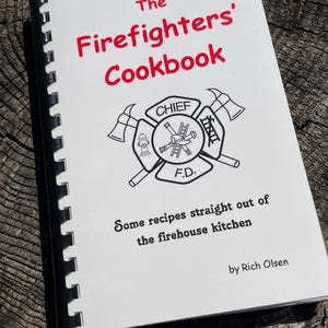 May include: A cookbook titled "The Firefighters' Cookbook" with a white cover and black spiral binding. The title is in red, and the cover features a firefighter emblem. The text "Some recipes straight out of the firehouse kitchen" is also visible.