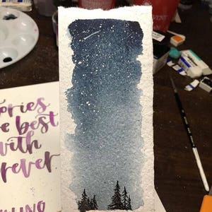 May include: A watercolor painting on handmade paper depicting a starry night sky with a shooting star. The sky is painted in shades of blue and white, with a gradient effect. The painting also features a row of small, dark trees in the foreground.