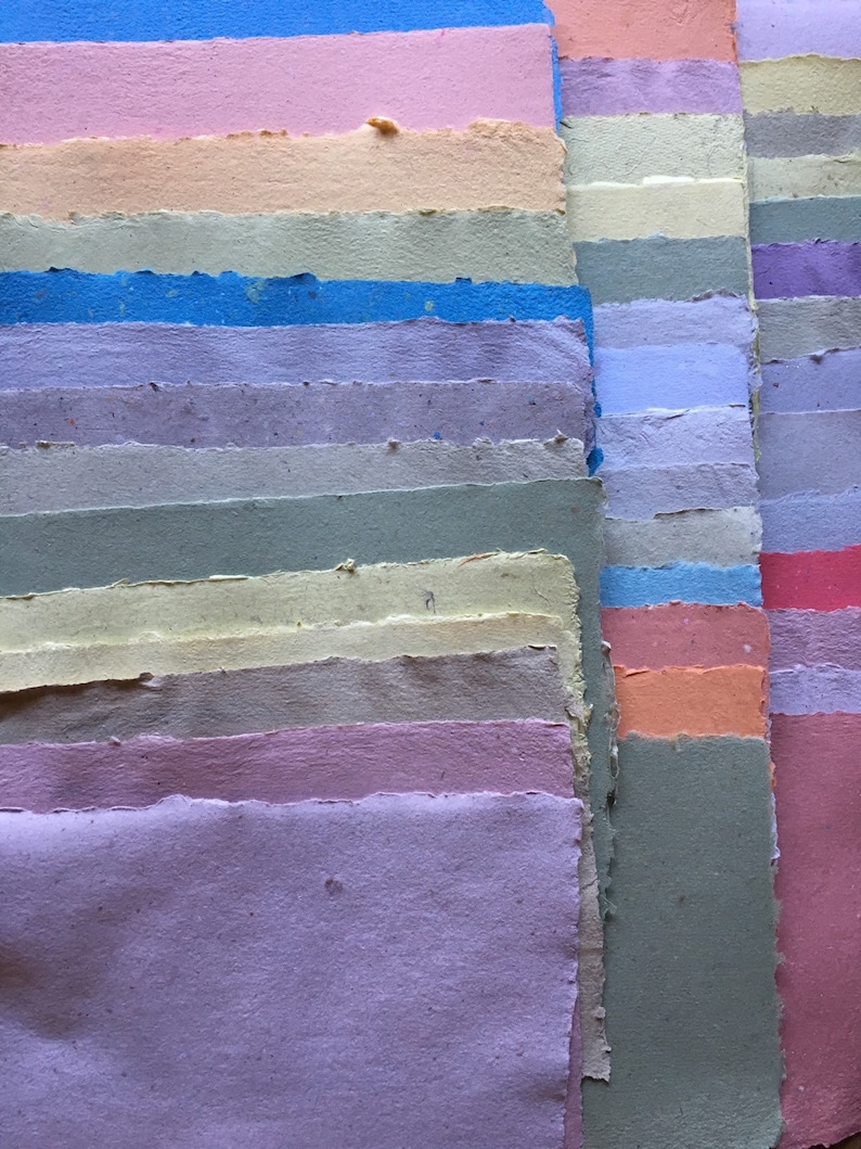 May include: A stack of handmade paper sheets in various colors, including blue, pink, peach, green, and purple. The paper has a textured, handmade look.